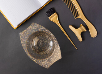 Golden hairdressers tools for hair coloring on a dark gray background . On the table are the accessories of the hair salon comb, brush, bowl and clips and notepad, top view.
