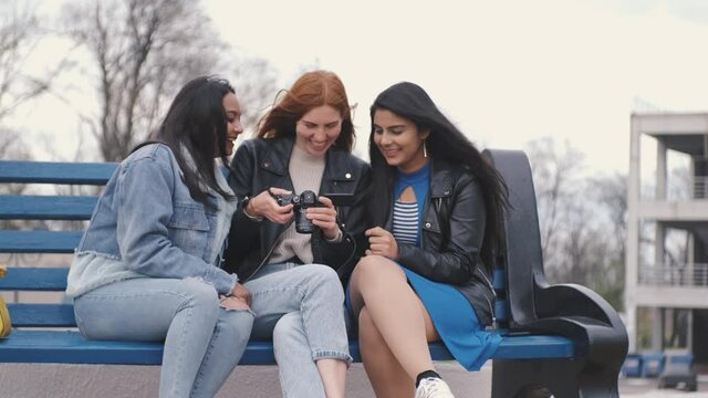 Redhaired caucasian photographer showing photos two beautiful longhaired indian women in public park, talking to each other, smiling and having fun