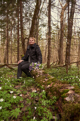 An elderly woman of 65 years with a bouquet of spring flowers sits on a fallen tree in the forest.