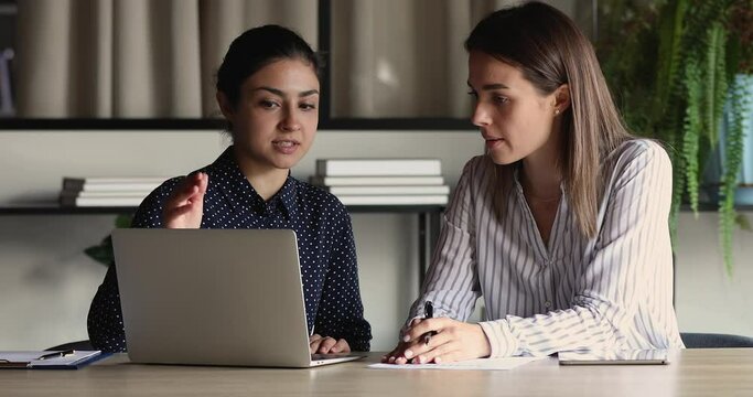 Focused millennial indian leader helping new worker with computer corporate software application in office. Motivated young mixed race employees developing project growth strategy, brainstorming ideas