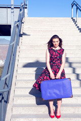 young woman in red dress and blue suitcase standing on a staircase at train station