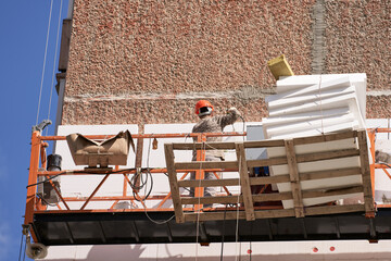 Naklejka premium Insulation and sound insulation of a residential building from the outside. A builder in a lifting cradle attaches Styrofoam slabs to the facade of a multi-storey building. Selective focus.