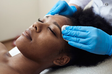 The cosmetologist cleanses with a tonic the face skin of a beautiful, young woman in a beauty...