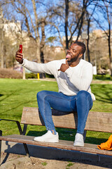 Young African American man sitting on a park bench, taking a self-portrait with a smartphone. Happy black man. New normality 