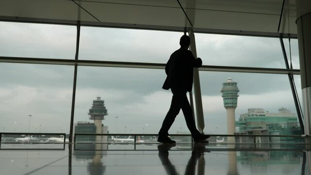 Silhouette of man standing against large window, looking to airport apron and buildings. Guy wait for boarding or take pause after arriving, long flight. He stay with arms on hips, wear casual jacket