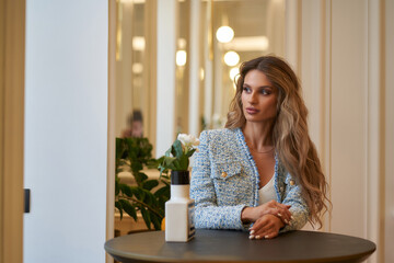 Beautiful caucasian woman with long wavy hair wearing blue tweed blazer sitting at table in bright...