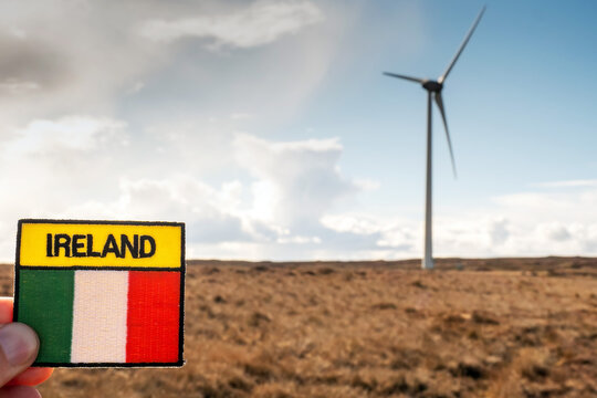 Badge with sign Ireland and Irish National flag in focus. Wind turbine out of focus. Renewable energy production in Ireland concept. Selective focus. Warm sunny day, cloudy blue sky.