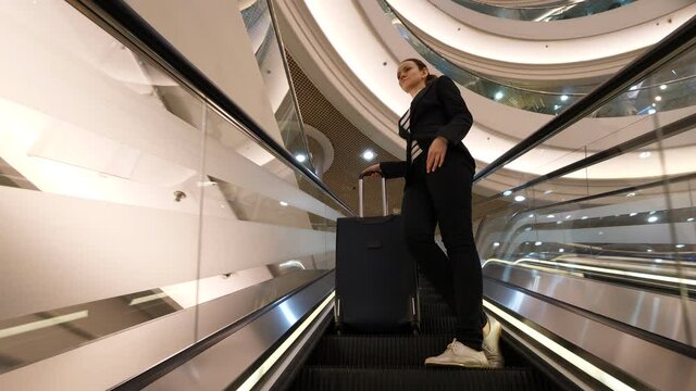 Traveller woman stay with large suitcase on escalator moving up, low angle shot. Modern shopping complex interior, large hall area. Business lady take free time to visit commercial centre