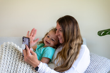 Young mom with daughter snuggled up on the couch using phone to video chat