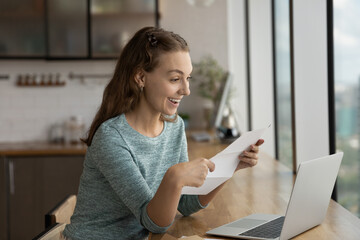 Excited young Caucasian woman work on laptop feel euphoric reading good news in paper letter in...