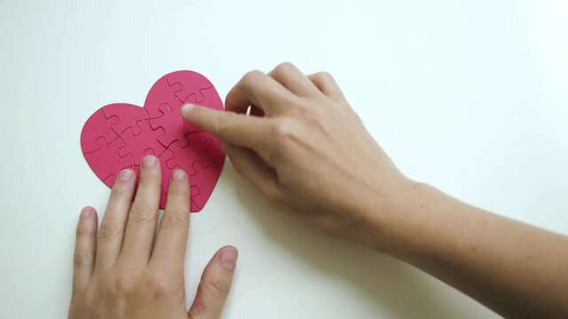Woman's hands places one piece of puzzle on the missing part to red heart shaped jigsaw on white table. Top view