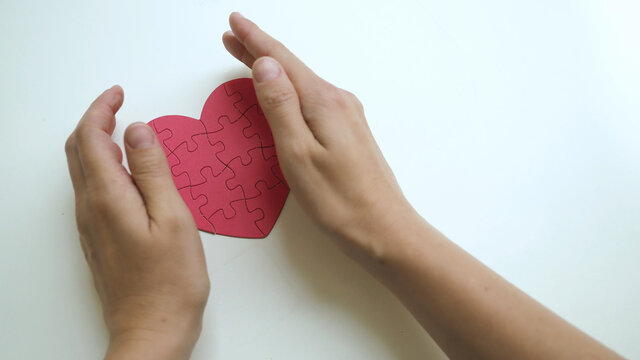 Top view on woman's both hands careful hug red heart shaped jigsaw from paper blocks puzzle on white table background