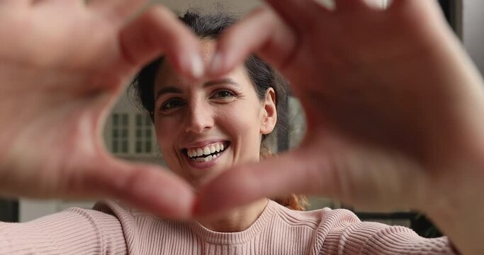Close up view face of lovely 30s happy smiling female vlogger gives love to followers, woman makes with fingers heart shape, symbol of devotion sign of sincere feelings. Volunteering, charity concept