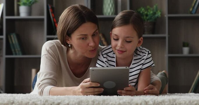 Close up loving mother and 7s daughter having fun use tablet, lying on soft carpet on warm floor with underfloor heat holding modern wireless device discuss purchase, enjoy educational website concept