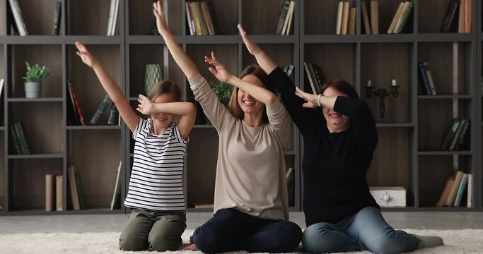 Happy three generations family 60s granny little girl her young mom sit on floor in modern living room make dab popular youth culture gesture have fun together at home, perform triumph dance concept