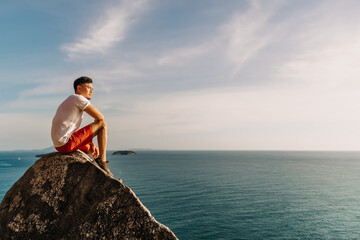 Fototapeta premium Asian man rest on the sea cliff rock after finish his hike trail.