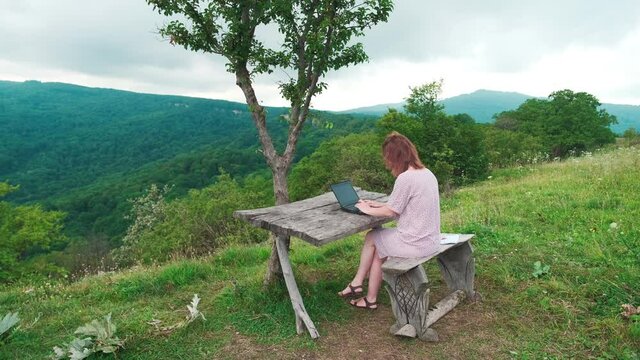 A girl with red hair works behind a laptop in nature, sits at a wooden table, around the mountain