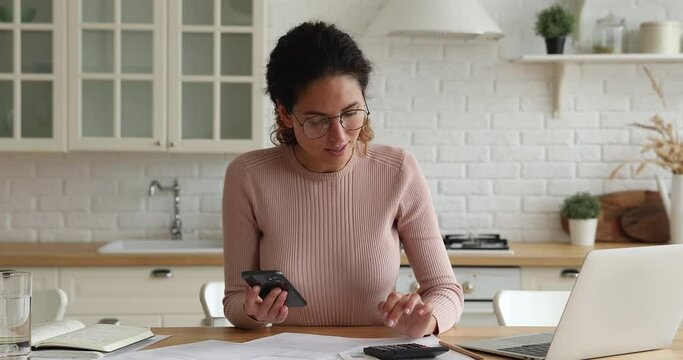 Young Accountant Woman In Glasses Use Calculator Count Expenses, Use Online Mobile E-bank App On Smartphone Smiles Feels Satisfied By Month Earnings Sale Result Take Notes Work Sit In Kitchen At Home