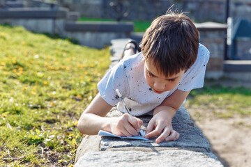 Cute boy writing something on a paper outside. Essay writing, college preparation concept