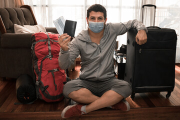 young adult man sitting on the wooden floor with his backpack and holding his travel suitcase with disposable masks to fight the coronavirus COVID-19