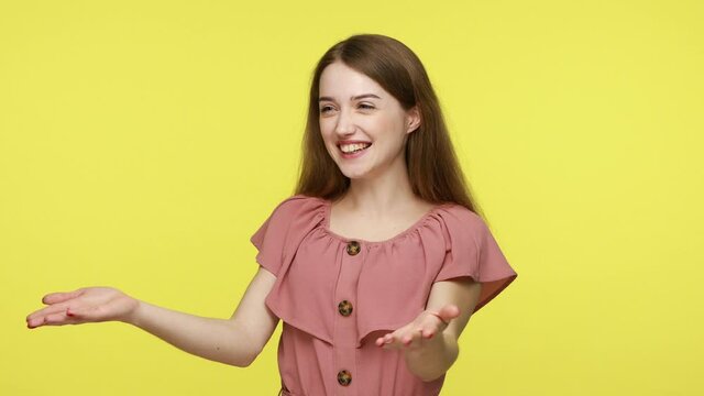 Amiable kind brown haired woman showing hand gesture take everything for free, sharing love care and looking at camera with charming smile. Indoor studio shot isolated on yellow background.
