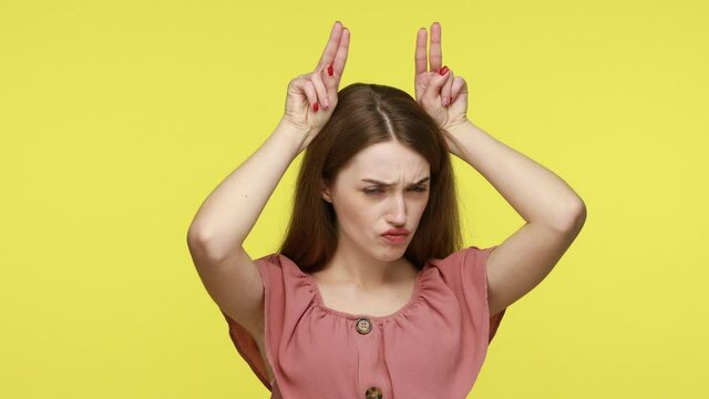 Playful cute girl with brown hair making bull horn gesture and puffing cheeks, pretending to be dangerous, woman showing she can attack, defence herself. Indoor shot isolated on yellow background.