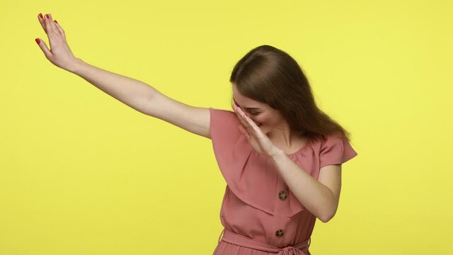 Attractive young woman with brown hair in dress performing dab dance with happy face and making dabbing movement with hands up, internet meme gesture. Indoor studio shot isolated on yellow background.