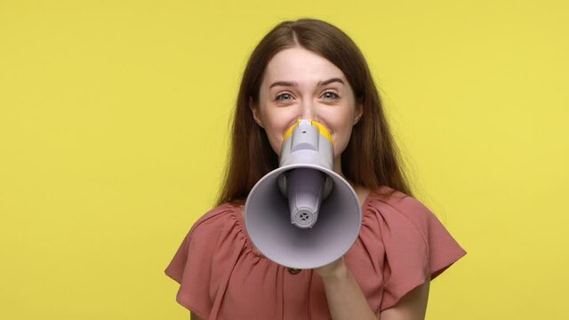 Closeup of happy young female wearing pink dress using loudspeaker for proclaiming breaking news or announcing advertisement, expresses positive emotions. Indoor shot isolated on yellow background.
