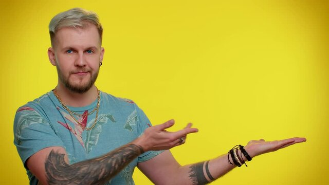 Handsome stylish trendy guy showing thumbs up and pointing empty place, advertising area for commercial text, copy space for goods promotion. Young adult man. Indoor studio shot on yellow background