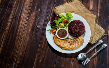 Top view flat lay shot of a sliced oven-roasted black pepper chicken with riceberry rice, sesame sauce, and salad side dish on the wooden table with spoon and fork. Concept of clean food, healthy