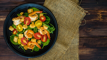 Top view flat lay shot of a homemade grilled shrimp salad with croutons, red and yellow cherry tomatoes in the black dish on the wooden table. Fresh prawn with vegetable. Concept of healthy food