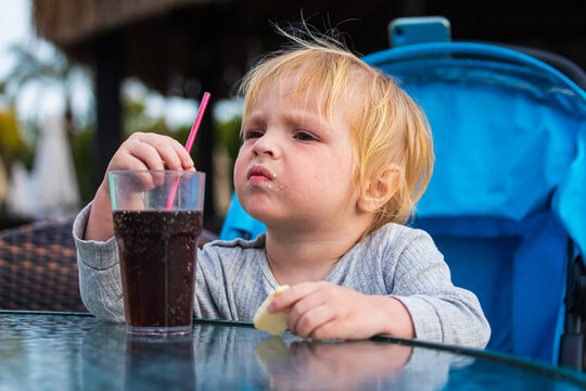 A Little Boy Is Sitting On A Chair And Happily Drinks Sparkling Water From A Glass Through A Straw. Feeding Process. The Baby Is Learning To Eat. Baby Food, Family, Child, Food