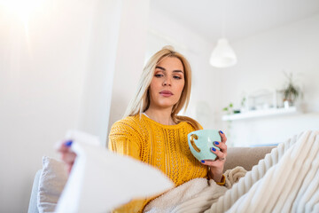 Sick attractive young woman has a cold and drinks hot tea upset young blonde in a sweater and blanket feels bad, cold, nausea, chills.