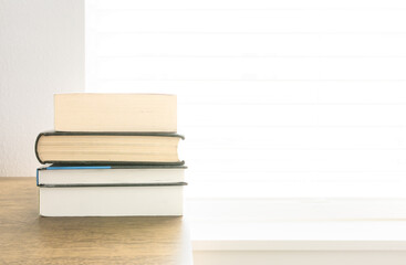 Many textbook, pocketbook stack on brown wooden nightstand table with bright natural sunlight thru window in bedroom, depth of field. Pile of books, student prepare for examination. Education concept.