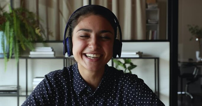 Head shot smiling pretty millennial indian ethnicity woman in headphones looking at camera, involved in distant video call zoom conversation, holding project negotiation remote meeting with colleagues