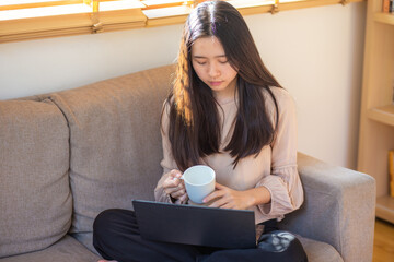 Woman sitting on sofa in an apartment drink coffee and opened laptop working at home