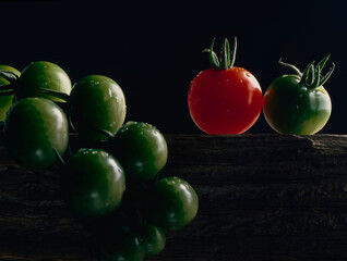 Bunch of green tomatoes and one red tomato on wooden plank against black background