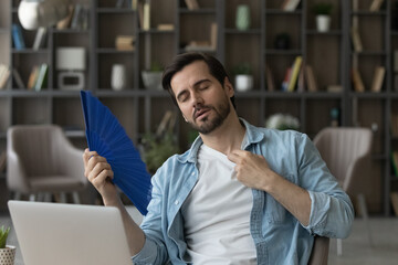 Exhausted businessman waving paper fan, sitting at desk with laptop, cooling, tired young man...
