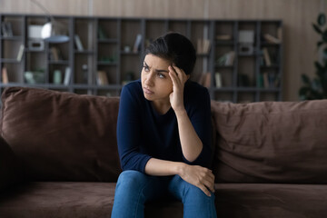 Thoughtful unhappy Indian woman lost in thoughts, looking to aside, sitting on couch at home alone,...