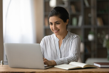 Smiling Indian woman using laptop, studying online at home, sitting at desk with books and...