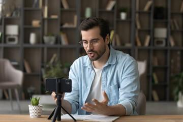 Young man blogger in glasses recording video, speaking, using smartphone standing on tripod,...