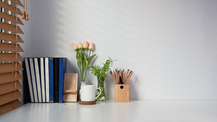 Contemporary workspace with book, coffee cup, pencil holder and flowers on white table.