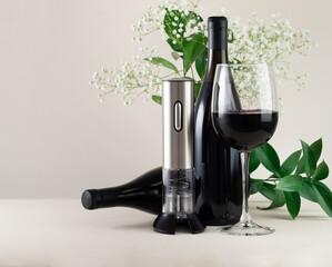 Electric corkscrew made of metal on a stand. Nearby are two bottles and a glass of wine. In the...