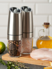 Electric spice grinders made of metal on a wooden cutting board. Chicken fillet next to it. White tile background. Close-up.