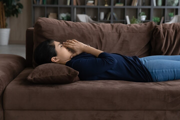 Obraz premium Side view unhappy Indian woman lying on couch alone, crying, thinking about personal problems, break up with boyfriend or divorce, unhealthy young female suffering from headache, feeling unwell