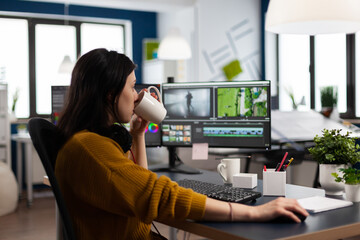 Professional colorist working in video footage using post production software, drinking coffee. Videographer editing audio film montage at pc with two monitors, sitting on desk in creative agency