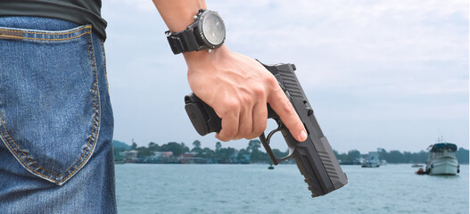 automatic black 9mm pistol holding in hand, ready to shoot, seascape background, concept for security, bodyguard, mafia, gangster, robbery and safety at the sea around the world.