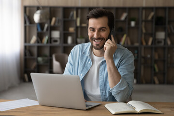 Head shot portrait smiling businessman talking on smartphone, using laptop, sitting at work desk, friendly manager freelancer consulting customer by phone call, negotiating, looking at camera