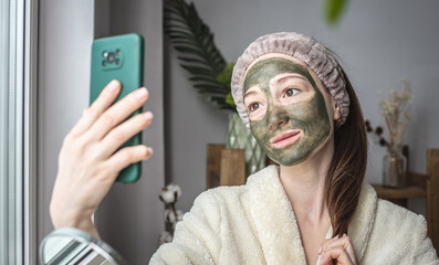 A young pretty woman in a bathrobe and with a green cosmetic mask on her face is taking a selfie on her mobile phone