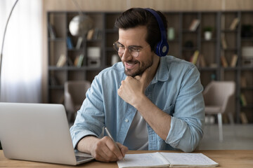 Close up smiling man in headphones and glasses using laptop, writing taking notes, motivated...